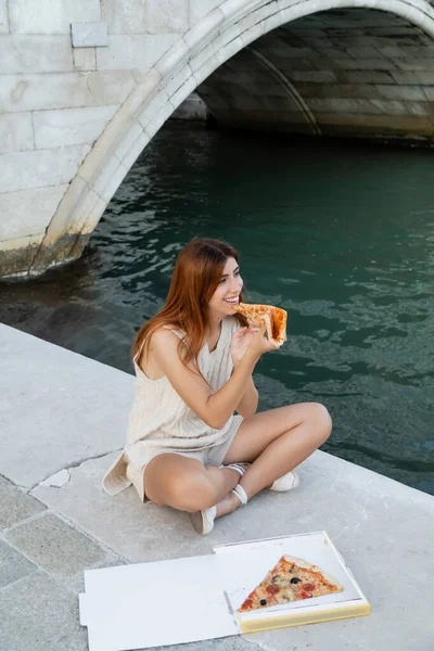 Mujer alegre sentada cerca del canal en Venecia y comiendo pizza tradicional - foto de stock