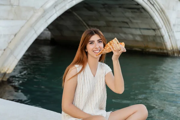 Cheerful woman with delicious pizza looking at camera near city canal in Venice — Stock Photo