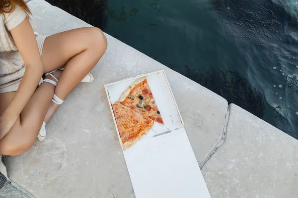 Зверху видніється смачна піца біля прокаженої жінки, яка сидить зі схрещеними ногами біля води у Венеції. — стокове фото