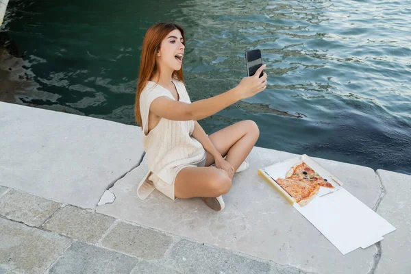 Aufgeregte Frau macht Selfie, während sie neben Karton mit leckerer Pizza auf Bürgersteig in Venedig sitzt — Stockfoto