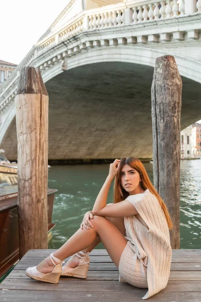 Rothaarige Frau in Strickkleidung und Keilsandalen sitzt auf einem Pier in Venedig und blickt in die Kamera — Stockfoto