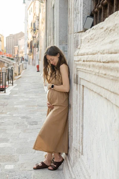 Smiling pregnant woman touching tummy near building on street in Venice — Stockfoto