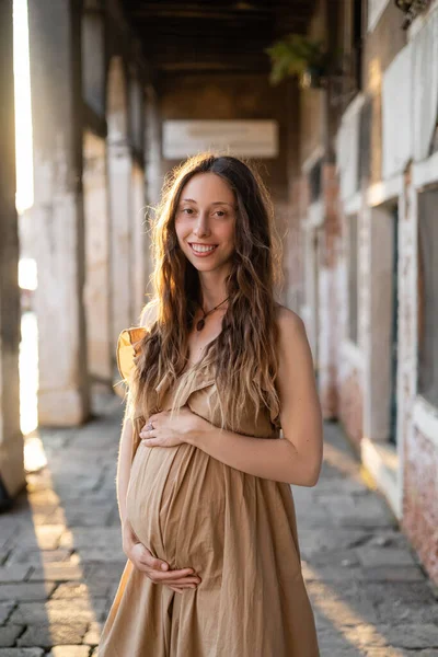 Femme enceinte positive en robe regardant la caméra près de vieux bâtiment flou en Italie — Photo de stock