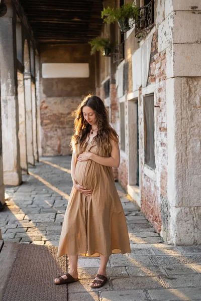 Mujer embarazada en vestido de verano tocando vientre en la calle urbana en Venecia - foto de stock