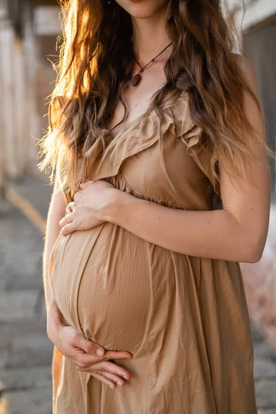 Vista cortada da mulher grávida tocando barriga enquanto está em pé à luz do sol na rua urbana — Fotografia de Stock