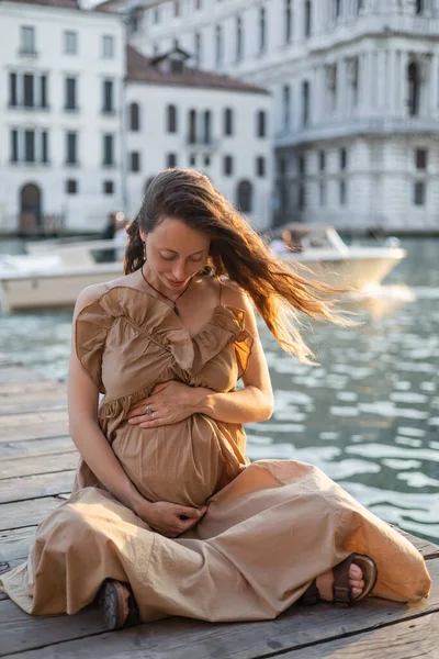 Smiling pregnant woman sitting on wooden pier in blurred Venice - foto de stock