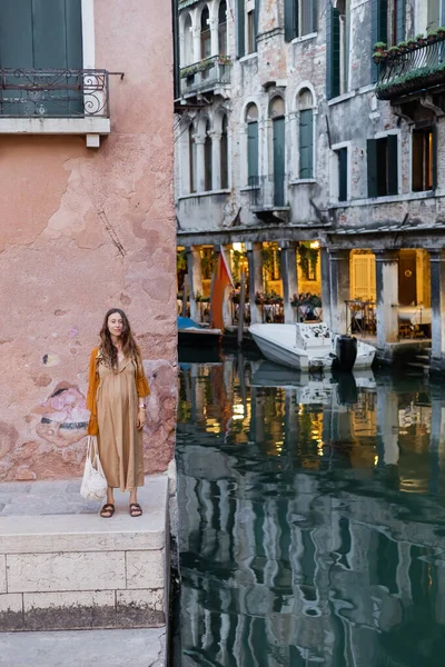 Улыбающаяся беременная женщина с веревочным мешком, стоящая возле здания и реки в Венеции — стоковое фото