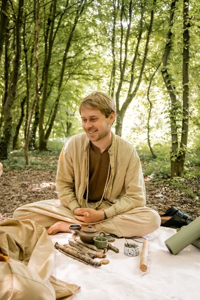 Улыбающийся мужчина сидит рядом с ароматной палочкой и чайником во время чайной церемонии в лесу — стоковое фото
