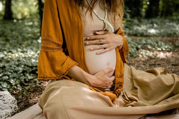 Обрізаний вид молодої вагітної жінки, яка торкається живота в лісі — стокове фото