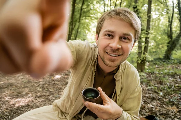 Uomo sorridente guardando la fotocamera mentre tiene la tazza di tè nella foresta — Foto stock