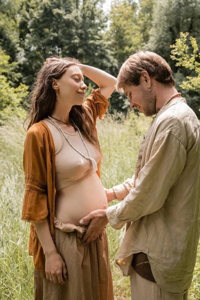 Side view of smiling man touching belly of pregnant wife in forest - foto de stock