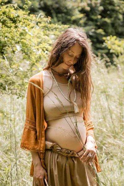 Brünette schwangere Frau hält Gras in der Nähe des Bauches im Wald — Stockfoto