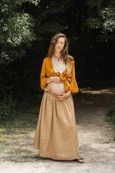 Молода вагітна жінка посміхається і торкається живота на шляху в лісі — стокове фото