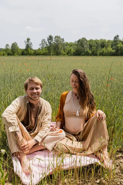 Homme souriant assis sur une couverture près d'une femme enceinte dans un champ — Photo de stock