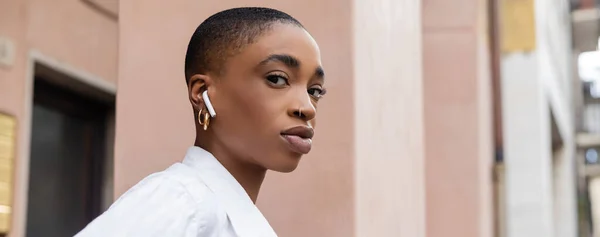 Femme afro-américaine à la mode avec des cheveux courts écoutant de la musique dans un écouteur dans la rue à Trévise, bannière — Photo de stock