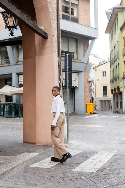 Stylish african american woman with wireless earphone looking at camera while walking on urban street in Treviso — Stock Photo