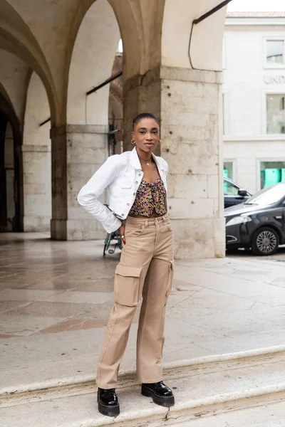 Fashionable african american woman standing near blurred old building on urban street in Treviso — Stock Photo