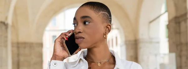 Mujer afroamericana de pelo corto hablando en un teléfono inteligente en una calle urbana borrosa en Treviso, pancarta - foto de stock