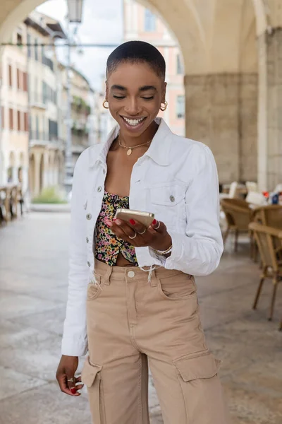 Femme afro-américaine souriante et élégante utilisant un smartphone dans la rue urbaine de Trévise — Photo de stock