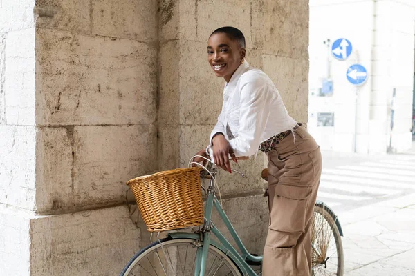 Allegro turista afroamericano dai capelli corti che guarda la macchina fotografica vicino alla bicicletta in strada in Italia — Foto stock