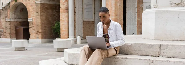 Fashionable african american blogger using laptop on stairs on street in Italy, banner — Stock Photo