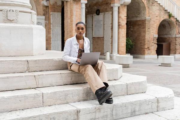 Short haired african american freelancer using laptop and looking at camera on stairs on urban street in Italy — Stock Photo