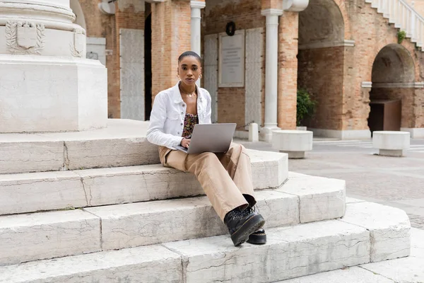 Freelance afro-americano dai capelli corti che utilizza laptop e guarda la fotocamera sulle scale in strada urbana in Italia — Foto stock