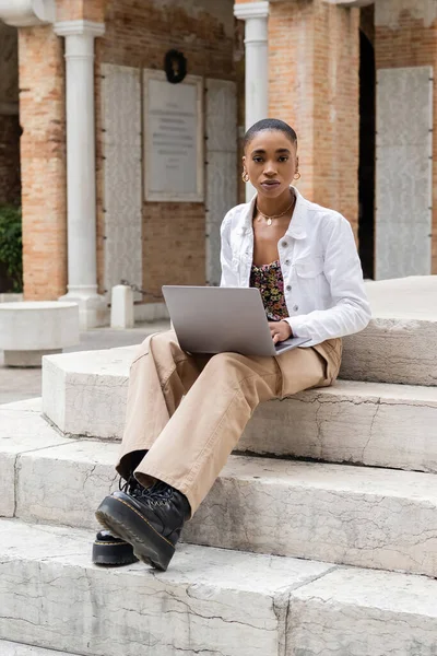 Trendy african american blogger with laptop looking at camera on stairs on urban street in Italy — Stock Photo
