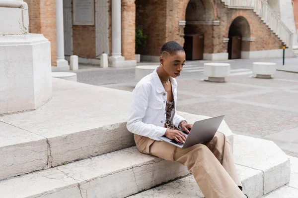 Trendy african american woman using laptop while sitting on stairs on urban street in Italy — Stock Photo
