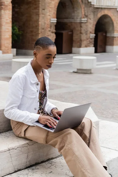 Stylish african american woman in white jacket using laptop on stairs on urban street in Italy — Stock Photo