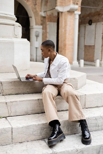 Vista lateral del freelancer afroamericano de pelo corto usando laptop en la calle urbana en Italia - foto de stock
