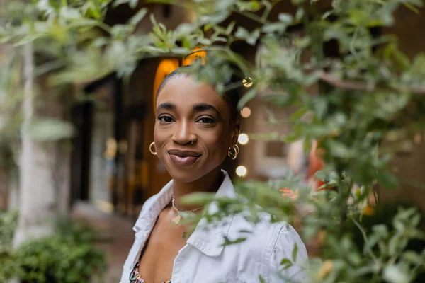 Souriante femme afro-américaine aux cheveux courts regardant une caméra près d'un arbre dans une rue urbaine — Photo de stock