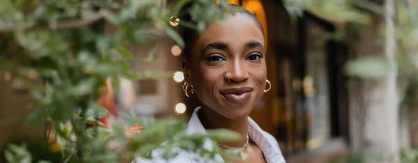 Stylish african american woman smiling at camera near tree on street, banner — Stock Photo