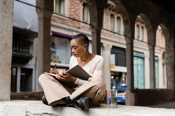 Mujer afroamericana de moda escribiendo en un cuaderno cerca de una botella de agua en la calle urbana de Treviso - foto de stock