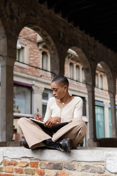 Mujer afroamericana de pelo corto escribiendo en cuaderno en la calle de la ciudad en Treviso - foto de stock