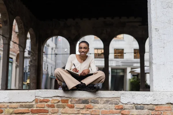 Веселые короткие волосы африканская женщина пишет на блокноте на открытом воздухе в Тревизо — стоковое фото