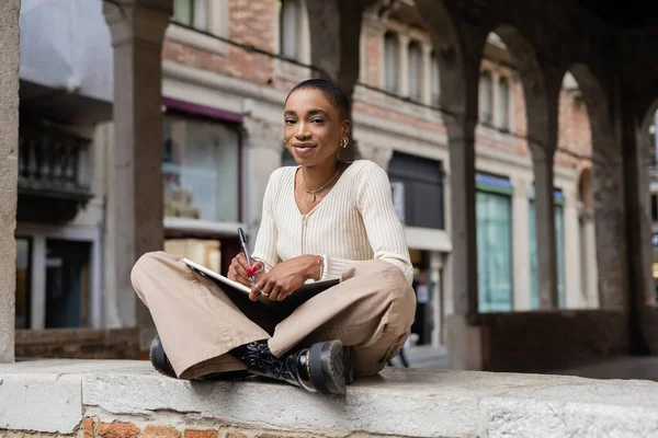 Stylish short haired african american woman writing on notebook near blurred building on urban street in Treviso — Stock Photo