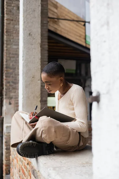 Femme afro-américaine à la mode écrivant sur un cahier en arche de bâtiment à l'extérieur à Trévise — Photo de stock
