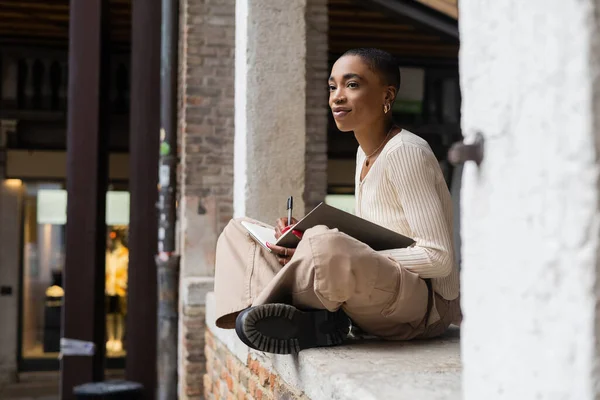 Stylish african american woman bolding pen and notebook in arch of building on street in Treviso — Stock Photo