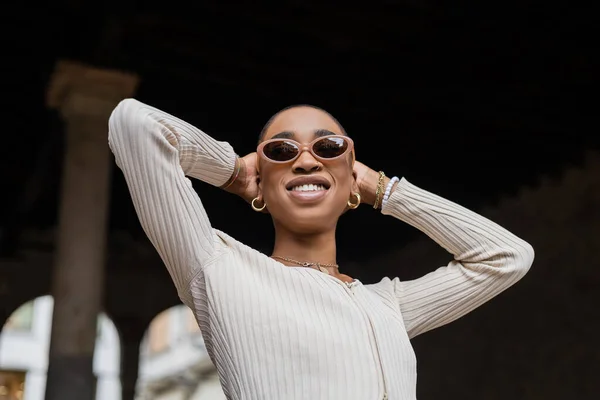 Vue à angle bas de souriante femme afro-américaine aux cheveux courts en lunettes de soleil debout à l'extérieur — Photo de stock