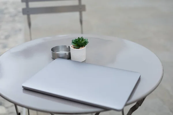 Ordenador portátil cerca de la planta y cenicero en la mesa del bistro al aire libre - foto de stock