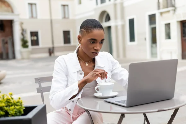 Стильная африканская американка смотрит на ноутбук рядом с кофе на городской улице в Италии — стоковое фото