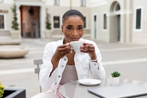 Elegante freelancer afroamericano que sostiene la taza de café cerca de la computadora portátil y planta en la cafetería al aire libre en Italia - foto de stock