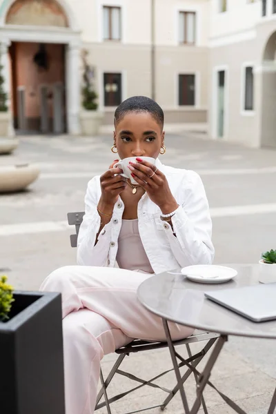 La mujer afroamericana a la moda que bebe el café cerca del portátil en la cafetería al aire libre en Italia - foto de stock