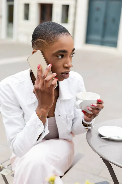 Stylish african american woman holding cup of coffee and talking on smartphone on urban street — Stock Photo