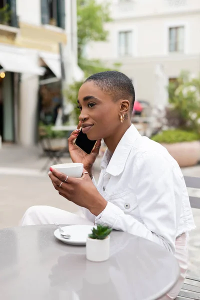 Stylish african american woman talking on cellphone and holding cup of coffee in outdoor cafe in Treviso — Stock Photo