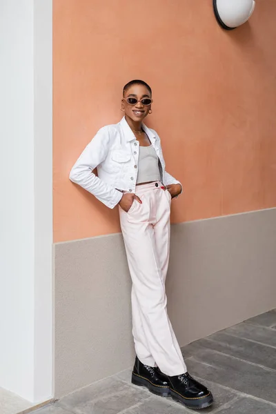 Femme afro-américaine élégante en lunettes de soleil souriant près du bâtiment à l'extérieur — Photo de stock