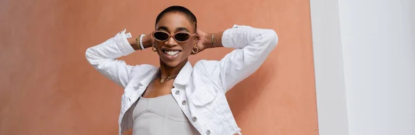 Femme afro-américaine aux cheveux courts en lunettes de soleil souriant près du mur à l'extérieur, bannière — Photo de stock