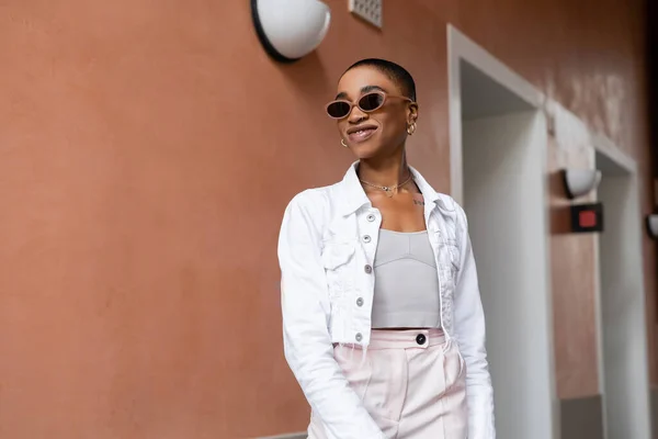 Sorridente donna afroamericana in occhiali da sole e giacca bianca in piedi sulla strada urbana — Foto stock