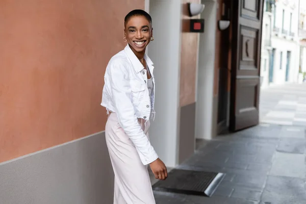 Fröhliche und stylische afrikanisch-amerikanische Frau blickt in die Kamera in der Nähe eines verschwommenen Gebäudes an der Stadtstraße in Treviso — Stockfoto